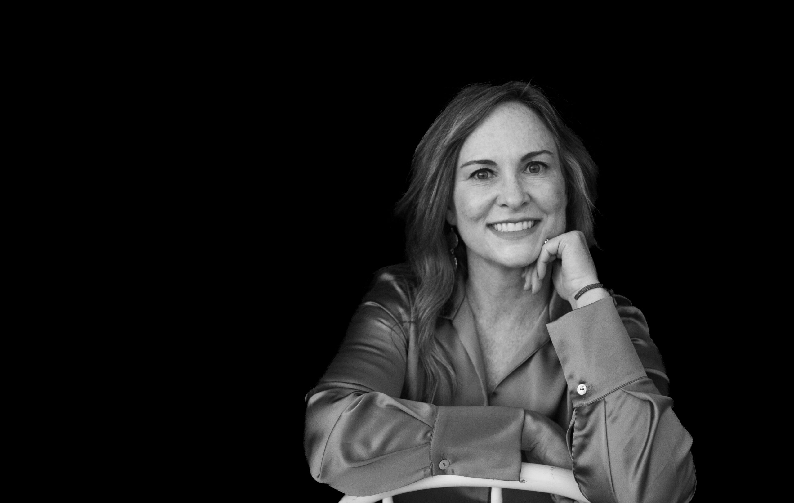 Woman looks at the camera while sitting backwards in a chair with her hand on her chin